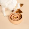 Een kleien hond staat gebogen boven een handgemaakte keramische slow feeder in een zalm roze kleur genaamd Blush. De voerbak is gevuld met gekleurde hondenbrokken van het merk PAWR. De hond heeft een witte vacht en bruine oren en draagt een waterbestendige halsband in beige en bruin met messing accenten gemaakt van BioThane. Zowel de halsband als de voerbak zijn van het merk RAWR pets