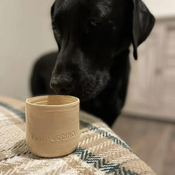 Zwarte labrador hond ruikt aan een puppuccino mok op een geruiten deken. De mok is handgemaakt en heeft een opdruk met PUPPUCCINO letters. De mok is naturel beige van kleur.